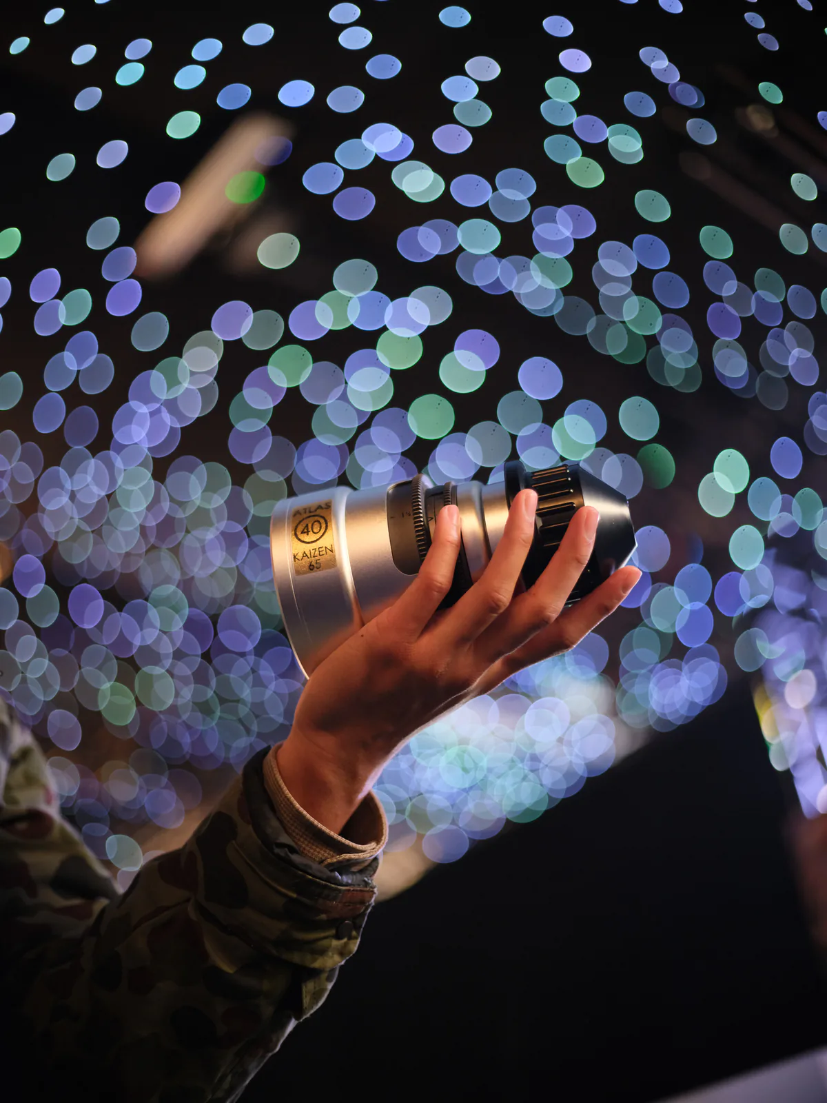 a hand holding an anamorphic cinema lens against blue and green bokeh